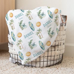 May include: A white baby blanket with a repeating pattern of surfboards, palm trees, waves, and suns in shades of blue, green, and yellow. The name "Daniel" is printed on the blanket. The blanket is folded in a black wire basket.