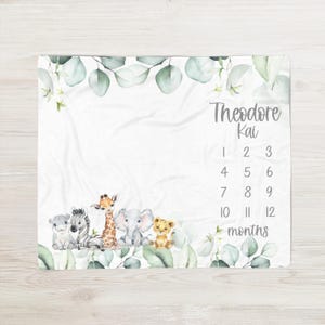 May include: A white baby milestone blanket featuring safari animal illustrations: a hippo, zebra, giraffe, elephant, and lion. The blanket includes eucalyptus leaves, the name "Theodore Kai", and numbers 1-12 for monthly photos.