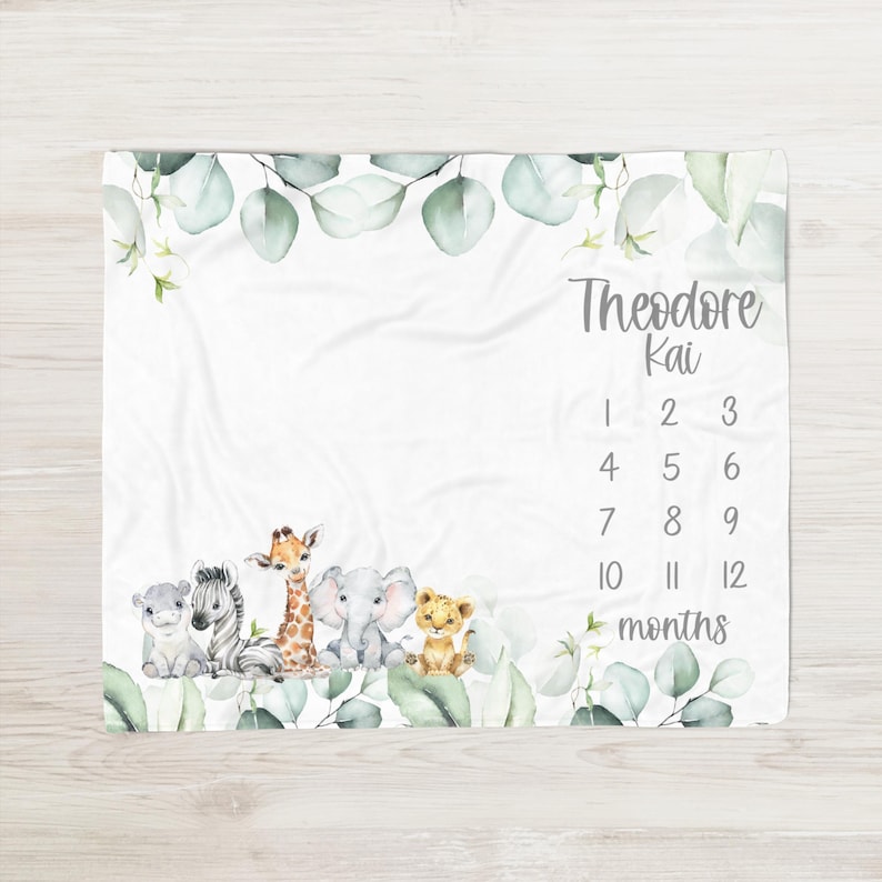 May include: A white baby milestone blanket featuring safari animal illustrations: a hippo, zebra, giraffe, elephant, and lion. The blanket includes eucalyptus leaves, the name "Theodore Kai", and numbers 1-12 for monthly photos.