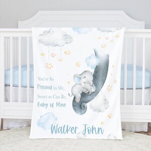 Pode incluir: Um cobertor de bebê branco com um design em aquarela com um elefante cinza, nuvens e estrelas. O cobertor tem o texto "You're So Precious to Me, Sweet as Can Be, Baby of Mine" e um nome em azul.
