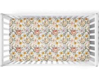 Puede incluir: Una sábana bajera floral para cuna con un patrón repetido de rosas, margaritas y otras flores en tonos melocotón, amarillo y crema. La sábana está dentro de una cuna blanca, ofreciendo una vista superior de la ropa de cama.
