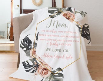 Può includere: Coperta in pile bianca con un design floreale e a foglie, e la parola "Mom" in elegante scrittura. La coperta presenta una cornice geometrica dorata con il testo "We love you".