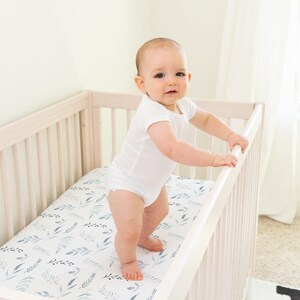 May include: A baby in a white onesie stands in a light wood crib. The crib mattress has a white background with a blue and gray leaf pattern. The crib is in a bright room with a white wall and a patterned rug.