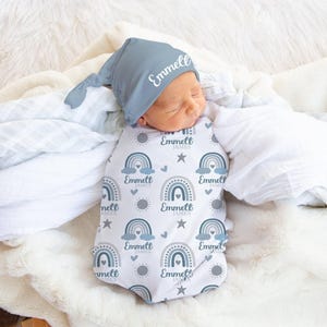 May include: A baby swaddled in a blanket and wearing a matching hat. The swaddle has a pattern of rainbows and stars in blue and grey, with the name "Emmett James" printed on it. The hat is a solid blue-grey with the name "Emmett" printed on it.
