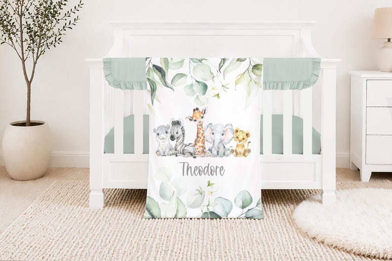 May include: A white cot with a mint green bumper and a matching blanket. The blanket features watercolor illustrations of a zebra, giraffe, elephant, lion, and other animals. The name "Theodore" is printed on the blanket.