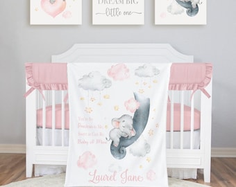 May include: A white baby crib with a pink and white bedding set. A white blanket with a watercolor elephant design, pink clouds, and the text "You're So Precious to Me, Sweet as Can Be, Baby of Mine" is draped over the crib. Three framed art pieces are above the crib.