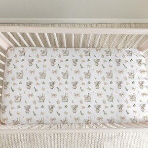 Peut inclure: Un lit bébé blanc avec un drap-housse à motifs répétés d'éléphants, de nœuds et d'éléments floraux gris et beige. Le lit est rose clair et posé sur un tapis crème.