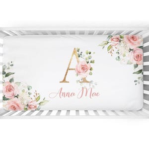 Könnte beinhalten: Ein weißes Kinderbettlaken mit einem Blumendesign mit rosa Rosen und grünem Laub. Das Laken hat einen goldenen Buchstaben "A" und den Namen "Anna Mae" in einer Schreibschrift. Das Laken befindet sich in einem weißen Kinderbett.