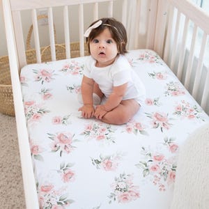 May include: A baby sits on a crib sheet with a floral pattern of pink roses and green leaves. The crib is made of light-colored wood. The baby is wearing a white onesie and a white bow in their hair.