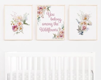 May include: Three framed floral art prints with watercolor flowers in pink, peach, and blue hues. One print features the text "You belong among the Wildflowers" in a script font. The prints are displayed above a white crib.