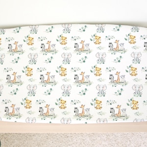 May include: A white changing mat cover featuring a repeating pattern of watercolour safari animals, including elephants, giraffes, lions, and zebras, with green foliage. The mat is on a light wood changing table.