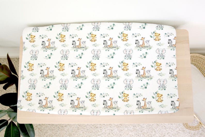 May include: A white changing mat cover featuring a repeating pattern of watercolour safari animals, including elephants, giraffes, lions, and zebras, with green foliage. The mat is on a light wood changing table.