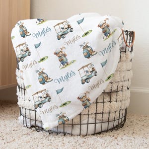 May include: White baby blanket featuring a repeating pattern of a teddy bear golfer, golf carts, golf bags, and flags. The name "Myles" is written in a script font. The blanket is folded in a wire basket.