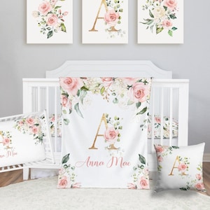 May include: A nursery set featuring a white crib, bedding, and decorative wall art. The set includes a crib sheet, blanket, and pillow, all adorned with pink roses, greenery, and the letter "A". The name "Anna Mae" is also displayed.