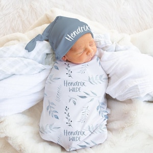 Puede incluir: Un bebé recién nacido envuelto en una manta blanca y azul con estampado de hojas y un gorro a juego. El gorro y la manta están personalizados con el nombre "Hendrix Wilde" en escritura blanca. El bebé está acurrucado sobre una manta blanca y suave.