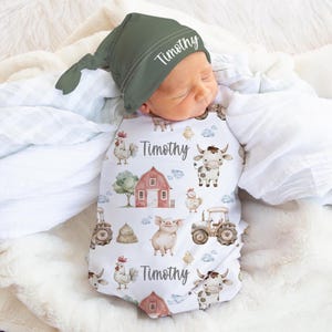 May include: A baby wrapped in a white swaddle blanket featuring a farm animal design with cows, chickens, and a tractor. The swaddle is printed with the name "Timothy". The baby wears a green knotted hat, also printed with the name "Timothy".