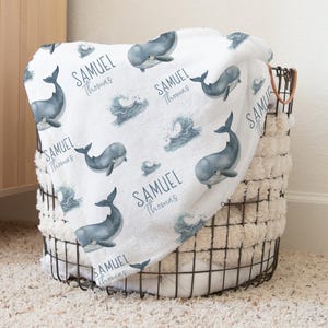 Könnte beinhalten: Eine weiße Babydecke mit Wal- und Wellenmuster, personalisiert mit dem Namen "Samuel Thomas". Die Decke ist über einem Drahtkorb mit einem flauschigen, cremefarbenen Futter drapiert.