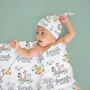 May include: A white baby blanket and matching hat with a repeating pattern of safari animals, including lions, giraffes, and elephants, with the name "Jonah DAVID" printed on them. The baby is wrapped in the blanket.