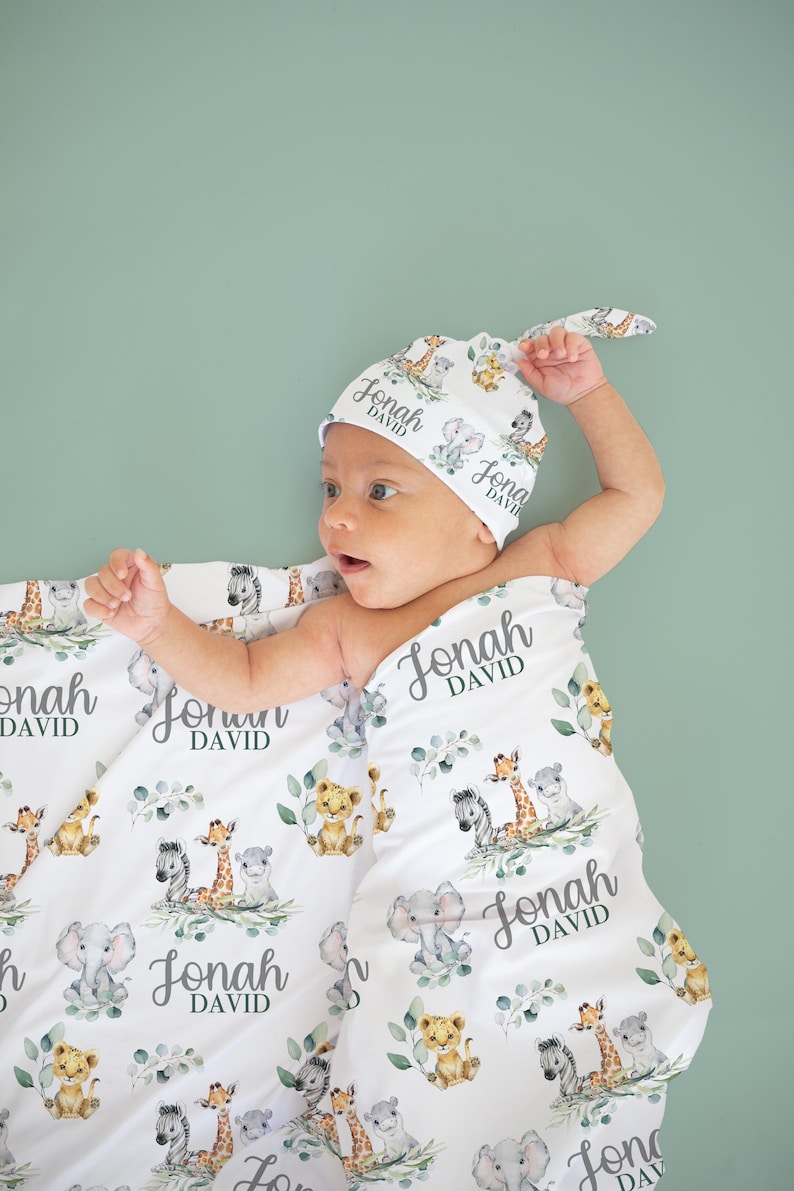 May include: A white baby blanket and matching hat with a repeating pattern of safari animals, including lions, giraffes, and elephants, with the name "Jonah DAVID" printed on them. The baby is wrapped in the blanket.