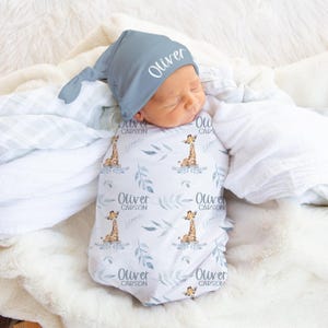 Puede incluir: Un bebé recién nacido envuelto en una manta envolvente blanca y azul con estampado de jirafas y un gorro azul a juego. El gorro tiene el nombre "Oliver" impreso. La manta presenta ilustraciones de jirafas y el nombre "Oliver Carson".