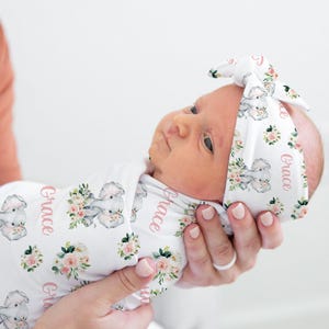 Puede incluir: Un bebé recién nacido envuelto en una manta blanca y con una diadema a juego. La manta y la diadema tienen un estampado floral y de elefantes con el nombre "Grace" en escritura rosa. El bebé está siendo sostenido.