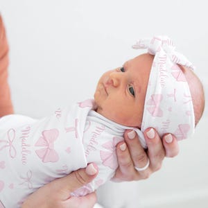 Peut inclure: Un nouveau-né enveloppé dans une couverture blanche et portant un bandeau assorti, tous deux ornés de nœuds roses et du nom "Madeline". Le bébé est tenu dans les bras de quelqu'un, sur un fond neutre.