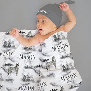 May include: A grey baby hat with the name "MASON" printed on it. The baby is wrapped in a white blanket featuring a wolf and forest design. The blanket also has the text "MASON Wolf" printed on it.