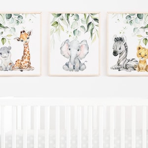 May include: Three framed watercolour prints. Each features a baby animal: a hippo and giraffe, an elephant, and a zebra with a lion cub. Green leaves border each print, set against a white background.