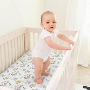 May include: A baby in a white onesie stands in a light-coloured wooden cot. The cot mattress has a white background with a pattern of animals and green foliage. The baby is holding onto the cot railing.