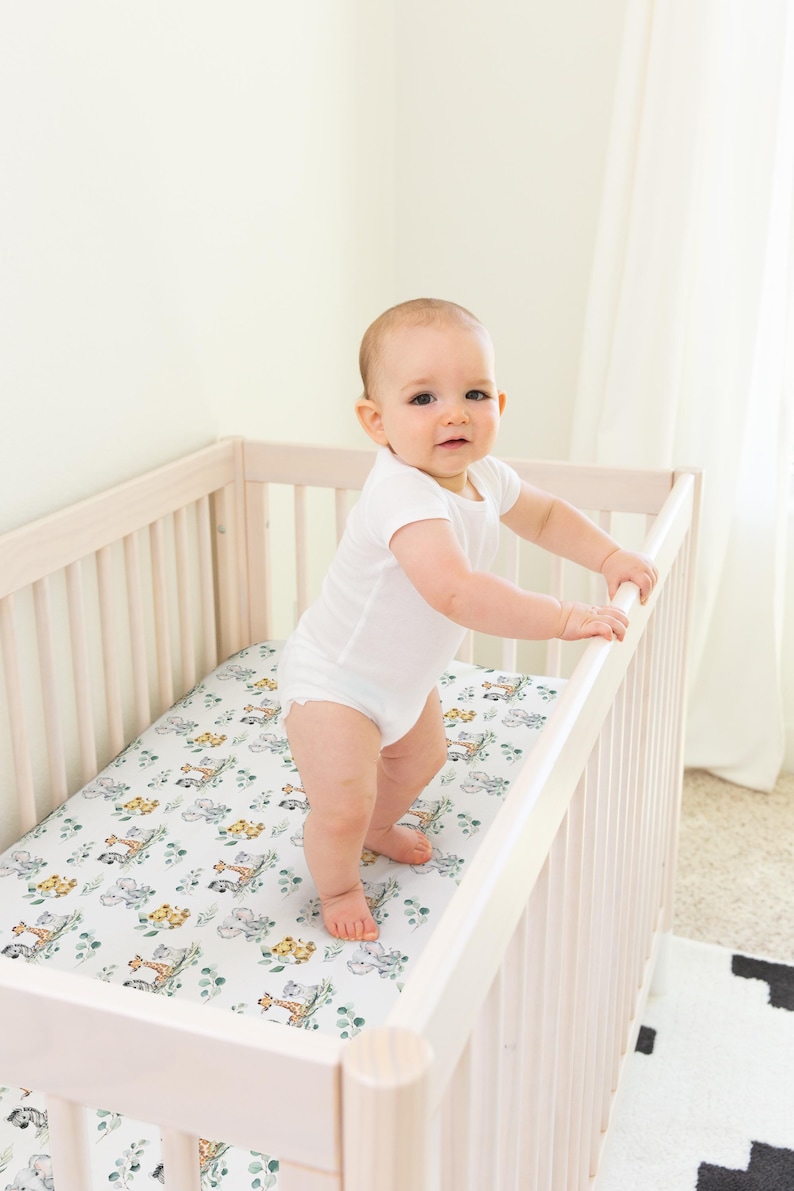 May include: A baby in a white onesie stands in a light-coloured wooden cot. The cot mattress has a white background with a pattern of animals and green foliage. The baby is holding onto the cot railing.