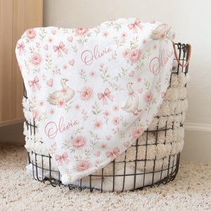 May include: A white baby blanket with a floral and goose pattern, featuring pink bows, roses, and hearts. The name "Olivia" is written in cursive. The blanket is draped over a wire basket with a fluffy, cream-colored lining.