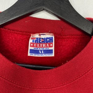 May include: A red sweatshirt with a white interior tag featuring the text TRENCH ULTRA, MADE IN THE U.S.A., XL, and 50% COTTON/50% POLYESTER. The garment has a ribbed crew neckline and is displayed on a black hanger against a white background.