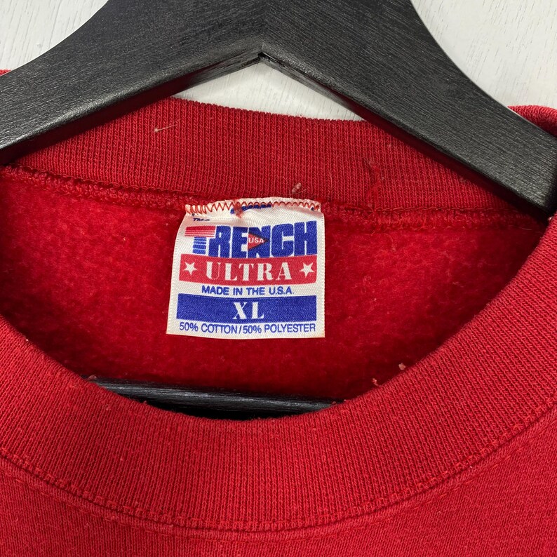 May include: A red sweatshirt with a white interior tag featuring the text TRENCH ULTRA, MADE IN THE U.S.A., XL, and 50% COTTON/50% POLYESTER. The garment has a ribbed crew neckline and is displayed on a black hanger against a white background.