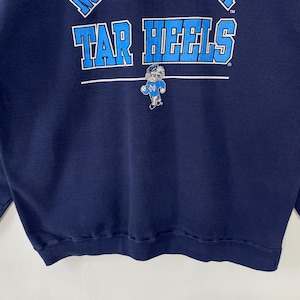 May include: Navy blue sweatshirt with the text "TAR HEELS" in light blue block letters across the chest. A small graphic of a Tar Heel mascot is below the text. The sweatshirt is made of a soft material.
