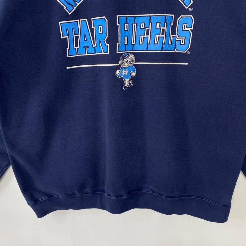 May include: Navy blue sweatshirt with the text "TAR HEELS" in light blue block letters across the chest. A small graphic of a Tar Heel mascot is below the text. The sweatshirt is made of a soft material.