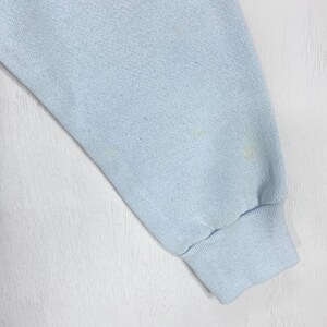 May include: A light blue long-sleeve garment sleeve featuring a ribbed cuff at the wrist. The fabric has a soft, textured appearance with subtle variations in color and a few faint, small yellowish spots visible on the surface of the material.