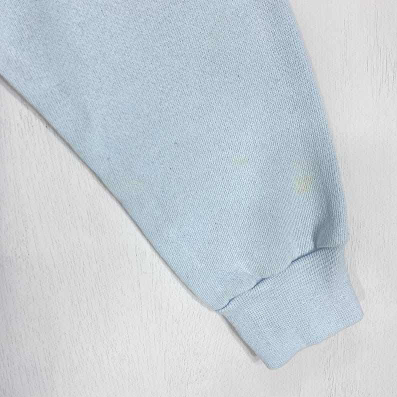 May include: A light blue long-sleeve garment sleeve featuring a ribbed cuff at the wrist. The fabric has a soft, textured appearance with subtle variations in color and a few faint, small yellowish spots visible on the surface of the material.