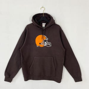 Puede incluir: Una sudadera con capucha de color marrón oscuro que presenta un gráfico de un casco de fútbol americano naranja con una franja blanca y una máscara facial blanca centrado en el pecho. Incluye un bolsillo tipo canguro y mangas largas.