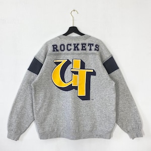 Könnte beinhalten: Graues Sweatshirt mit dem Wort "ROCKETS" über den Schultern. Auf der Rückseite befindet sich eine große gelbe und marineblaue Grafik. Die Ärmel haben marineblaue Akzente. Das Sweatshirt hängt an einem schwarzen Kleiderbügel.