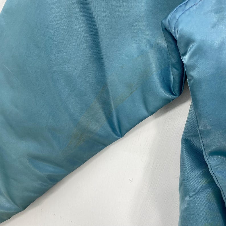 May include: Close-up of a light blue satin jacket. The fabric has a slight sheen and shows some discoloration. The jacket's seams are visible, and the overall texture is smooth.