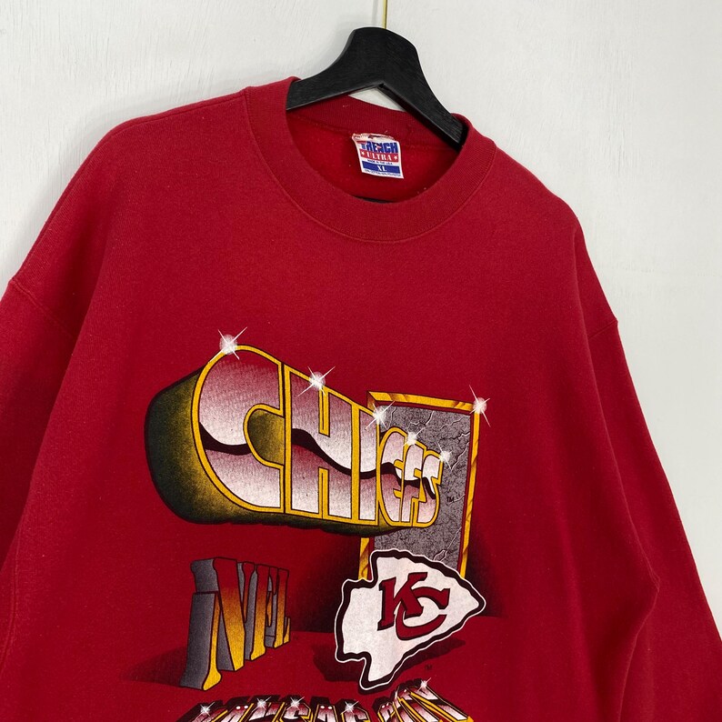 May include: A red crewneck sweatshirt featuring a graphic print with the text CHIEFS in a stylized font, the NFL logo, and the Kansas City Chiefs arrowhead emblem. The design includes sparkling star accents and a rectangular border element.