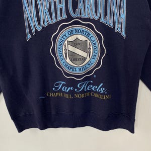 K&ouml;nnte beinhalten: Marineblaues Sweatshirt mit dem Schriftzug "NORTH CAROLINA" in Hellblau, der sich oben w&ouml;lbt. Darunter befindet sich ein rundes Emblem mit der Aufschrift "UNIVERSITY OF NORTH CAROLINA CHAPEL HILL, N.C." mit "LIBERTAS" und "LVX". Der Text "Tar Heels" ist darunter.