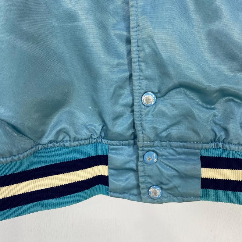 May include: Close-up of a light blue satin jacket. The jacket features a button-up front with three decorative buttons. The ribbed hem and cuffs have stripes in light blue, navy, and cream colors. The jacket has a shiny, smooth texture.