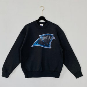 Puede incluir: Sudadera negra de cuello redondo con el logotipo de los Carolina Panthers. El logotipo es una cabeza de pantera estilizada en azul, negro y blanco. La sudadera está colgada de una percha negra sobre un fondo blanco.