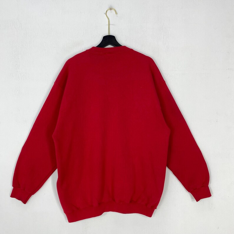 May include: A solid red long-sleeve sweatshirt featuring a crew neckline, ribbed cuffs, and a ribbed hemline. The garment is displayed hanging on a black plastic hanger against a plain white background, showcasing a simple and uniform design.