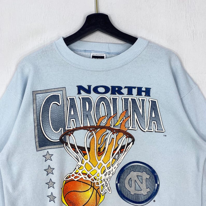 May include: A light blue crewneck sweatshirt featuring a graphic of a basketball going through a hoop with flames. The text reads NORTH CAROLINA in bold letters. Below the text are four grey stars and a circular logo with the letters NC.