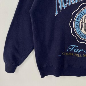 K&ouml;nnte beinhalten: Marineblaues Sweatshirt mit hellblauem und wei&szlig;em Universit&auml;tswappen und Text. Der Text enth&auml;lt "North Carolina University Chapel Hill, NC". Das Sweatshirt hat lange &Auml;rmel und einen Rundhalsausschnitt.