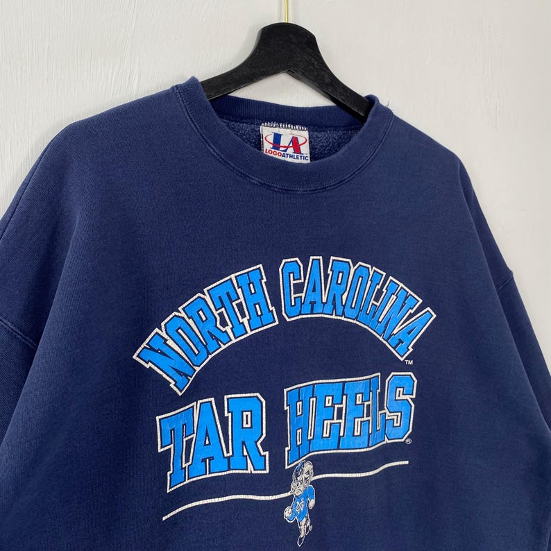 May include: Navy blue sweatshirt with the words "North Carolina Tar Heels" in white and light blue. The sweatshirt has a crew neck and long sleeves. The logo of the brand "Logo Athletic" is visible.