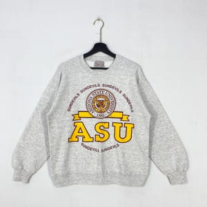 May include: A heather gray ASU Sundevils sweatshirt. The sweatshirt features a circular emblem with the words "Arizona State University" and "1885". Below the emblem is the word "ASU" in large gold letters.