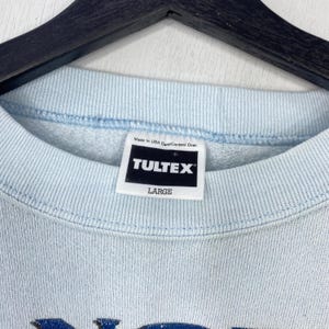 May include: A light blue crewneck sweatshirt featuring a white interior tag with black text that reads TULTEX and LARGE. The garment is displayed on a black hanger, showing the ribbed collar and the brand label positioned at the center neckline.